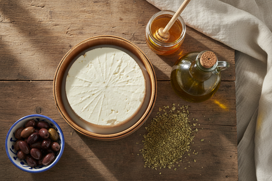 Authentic Greek pantry staples — barrel-aged PDO feta, extra virgin olive oil, thyme honey, and dried oregano arranged on a rustic wooden surface