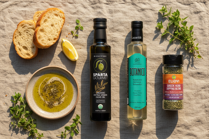 Sparta Gourmet Organic EVOO, Botanico White Balsamic, and Eliovi Greek Herb Dipping Blend on marble counter with ceramic dipping plate, crusty bread, and fresh herbs